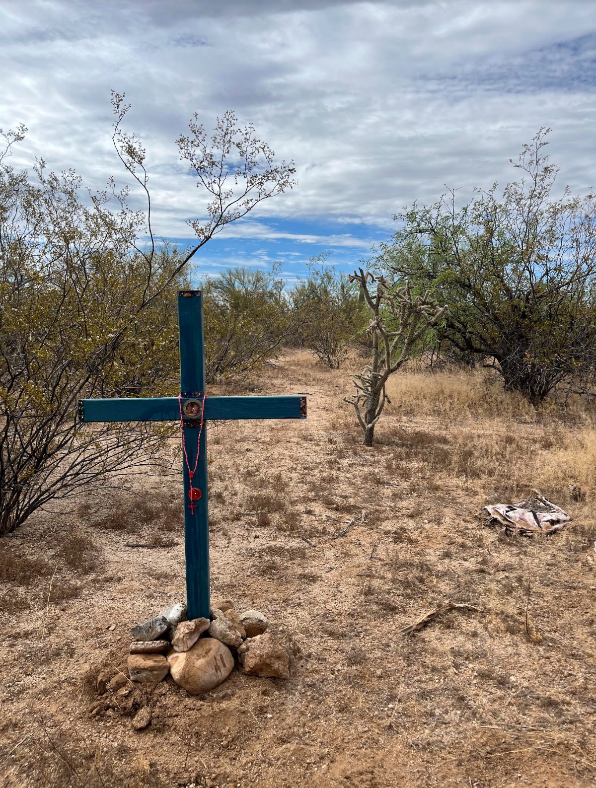 A homemade cross stands in the ground in a desert area surrounded by rocks and cacti. The location is in Southern Arizona near the U.S. - Mexico border.